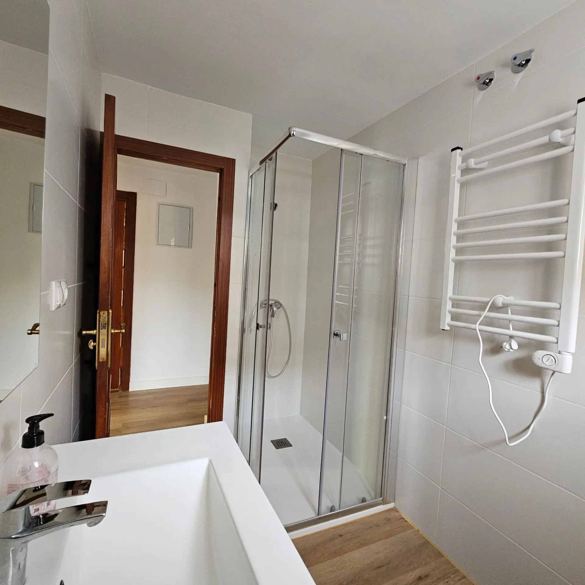 Baño reformado con ducha de mampara de cristal, radiador toallero blanco y suelo de madera